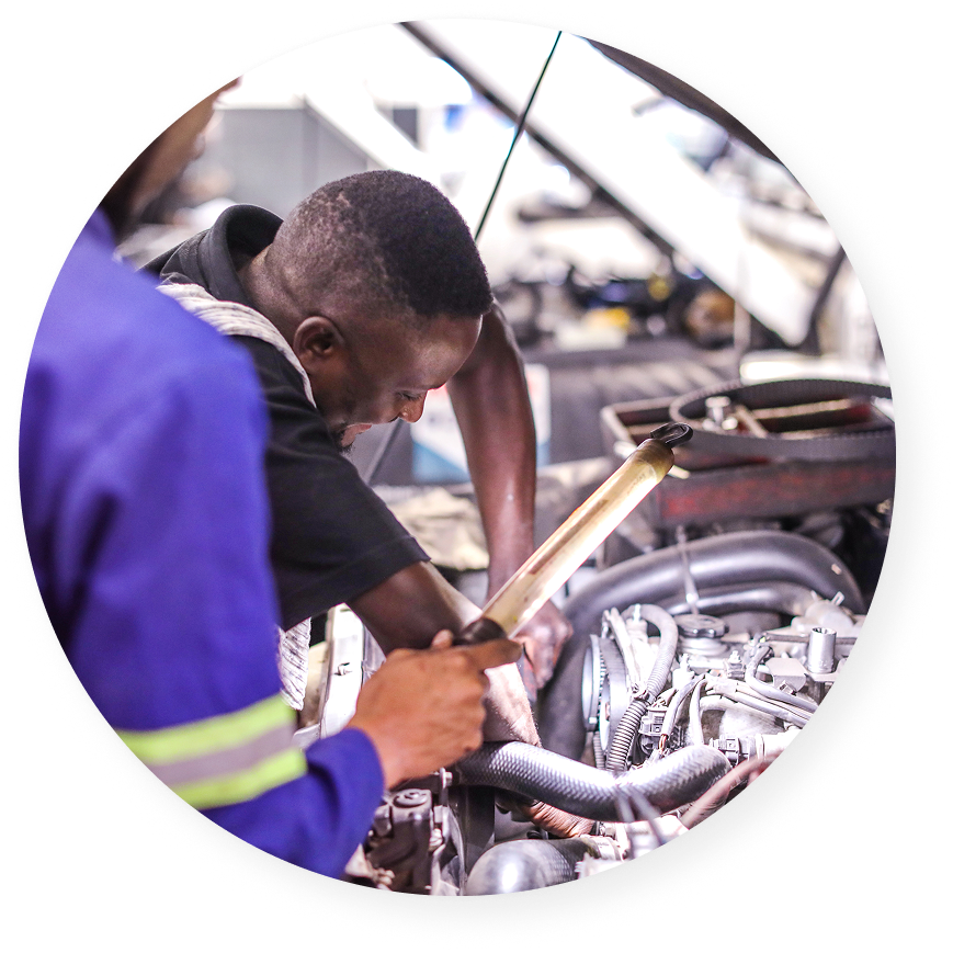 Two men working under the hood of a car in a mechanic workshop, one using a flashlight to illuminate the engine area.