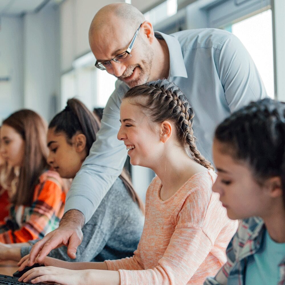 Teacher assisting a smiling student at a computer in a classroom while other students work nearby.