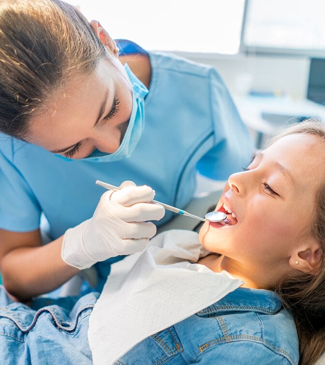 Dental hygienist wearing a mask and gloves examining a young girl’s teeth during a dental checkup.