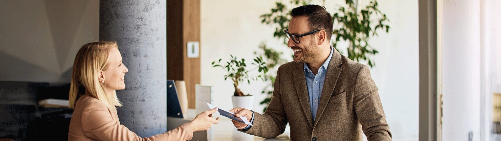 A hotel front desk staff member smiles while handing a key card or document to a guest at a modern reception area.