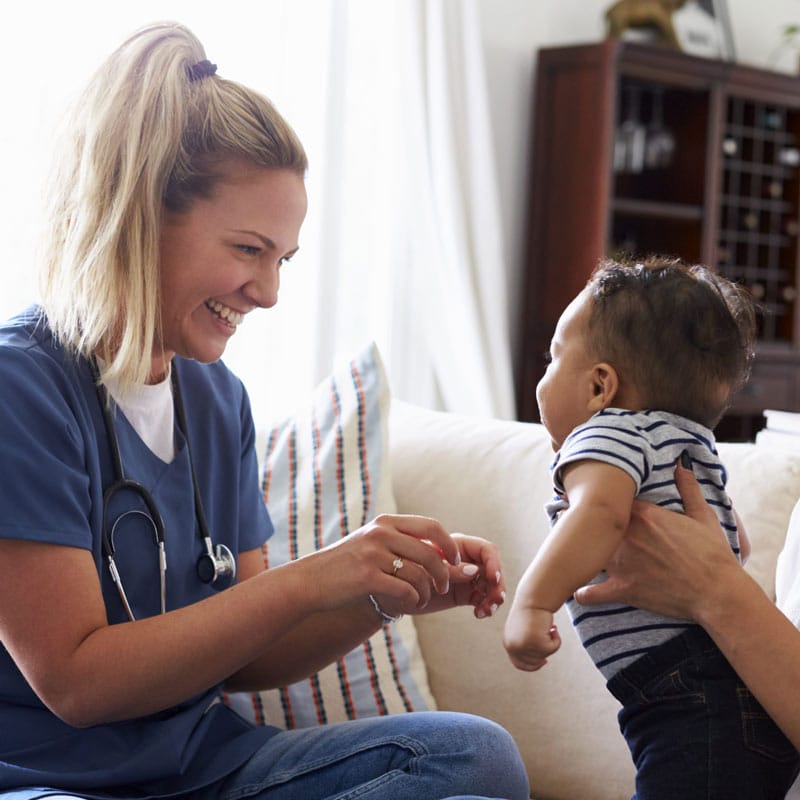 Nurse in blue scrubs smiling while interacting with a baby during a pediatric checkup in a home setting.
