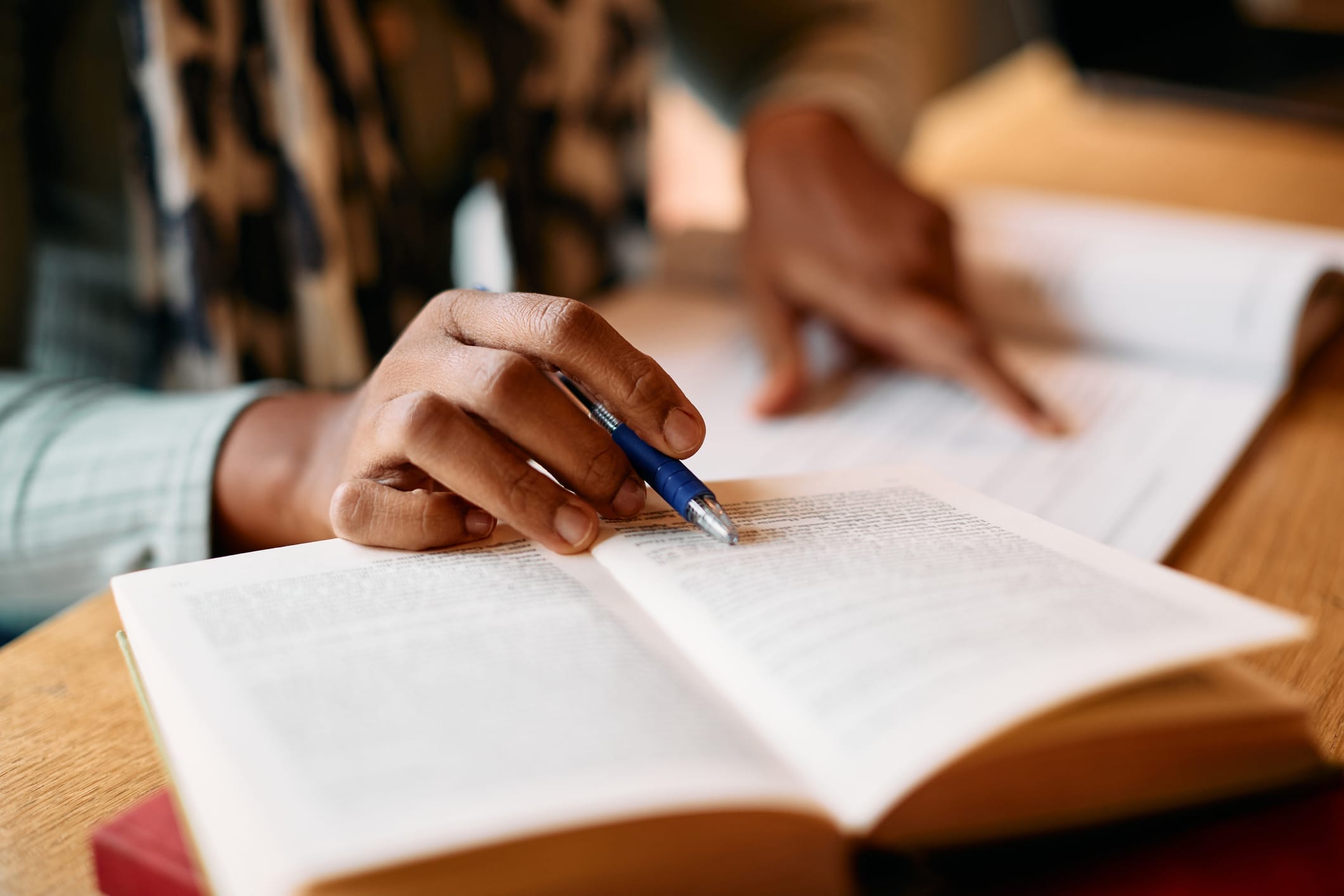 Close-up of a student’s hands holding a pen and reading an open textbook on a desk.