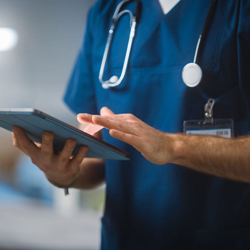 Healthcare professional in blue scrubs uses a tablet, with a stethoscope visible around their neck.