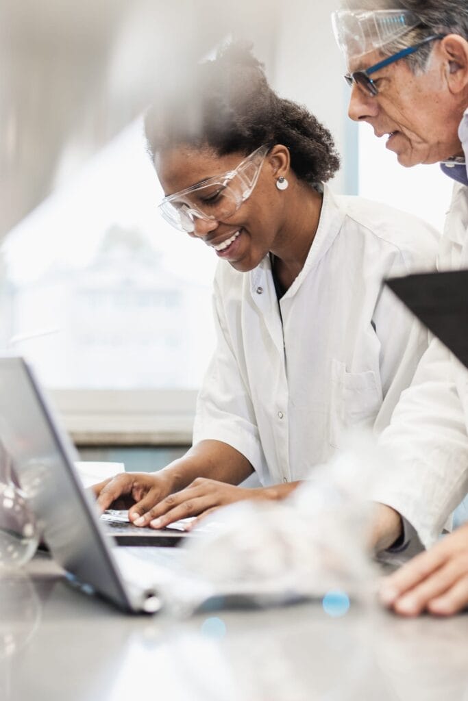 Student wearing safety goggles and a lab coat works on a laptop during a science lab while an instructor looks on nearby.