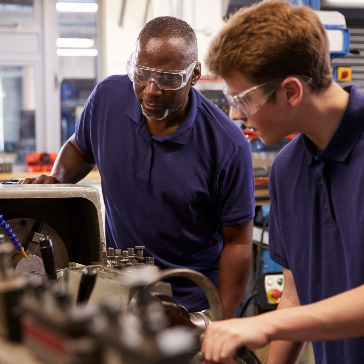 Adult instructor and student wearing safety goggles while operating machinery in a technical training workshop.