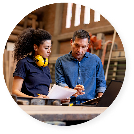 Young woman with safety earmuffs around her neck reviews papers with a mentor in a woodshop, both focused on a laptop screen.