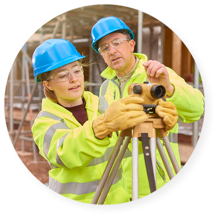 Two construction workers in safety gear adjust a surveying instrument on a tripod at an active job site.