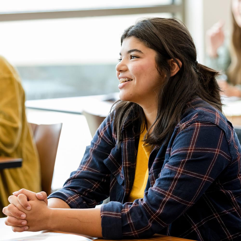 Smiling student seated at a classroom desk, listening attentively during a discussion with other students in the background.