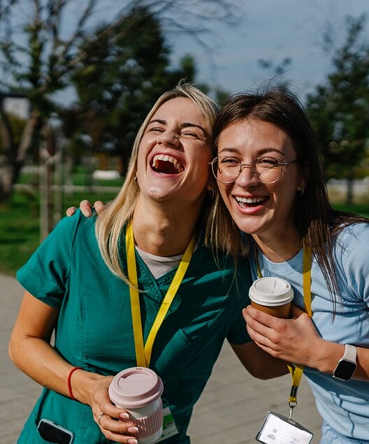 Two women in scrubs laugh together outdoors while holding coffee cups and wearing ID lanyards, standing on a campus walkway with trees in the background.