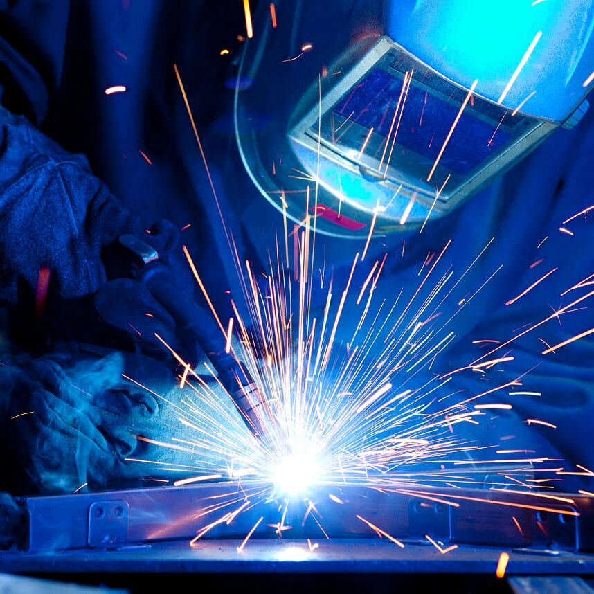 Welder wearing a protective helmet and gloves welding metal, with bright sparks flying from the welding torch.