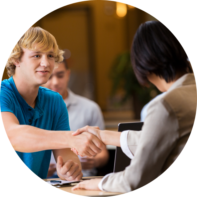 Young man in a blue shirt shaking hands with a woman across a table during a professional meeting, with another person seated in the background.